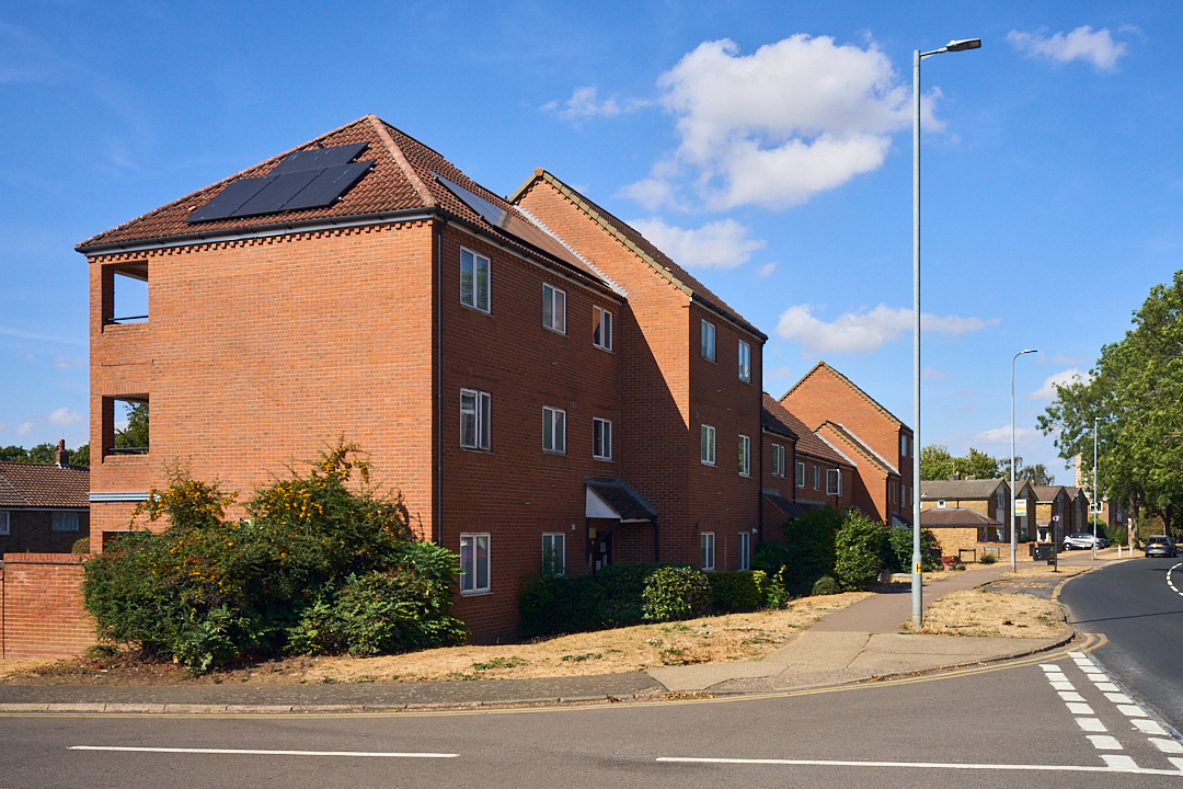 Street-level perspective of a brick social housing block in Stevenage with dark solar panels installed on its pitched roof under a bright blue sky.