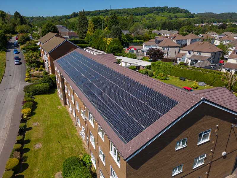 An aerial view of a long building with a brown roof covered in solar panels, situated in a green residential neighborhood next to a road.