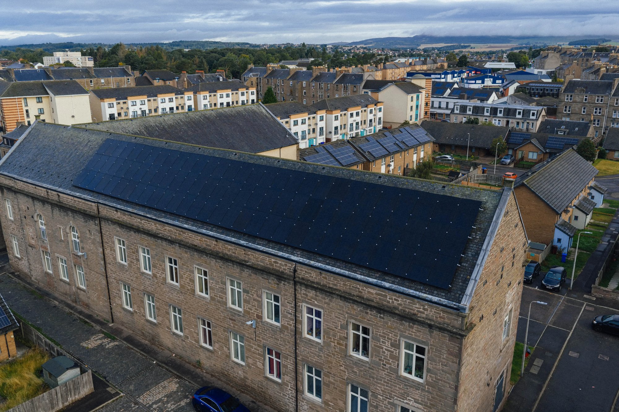 Aerial view of a large stone multi-family apartment building featuring a massive shared solar panel array installed across its long pitched roof.