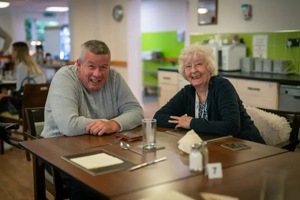 Two social housing residents at Willowbrook in Wales: a man and an older woman seated together at a dining table in a community room, smiling warmly at the camera.