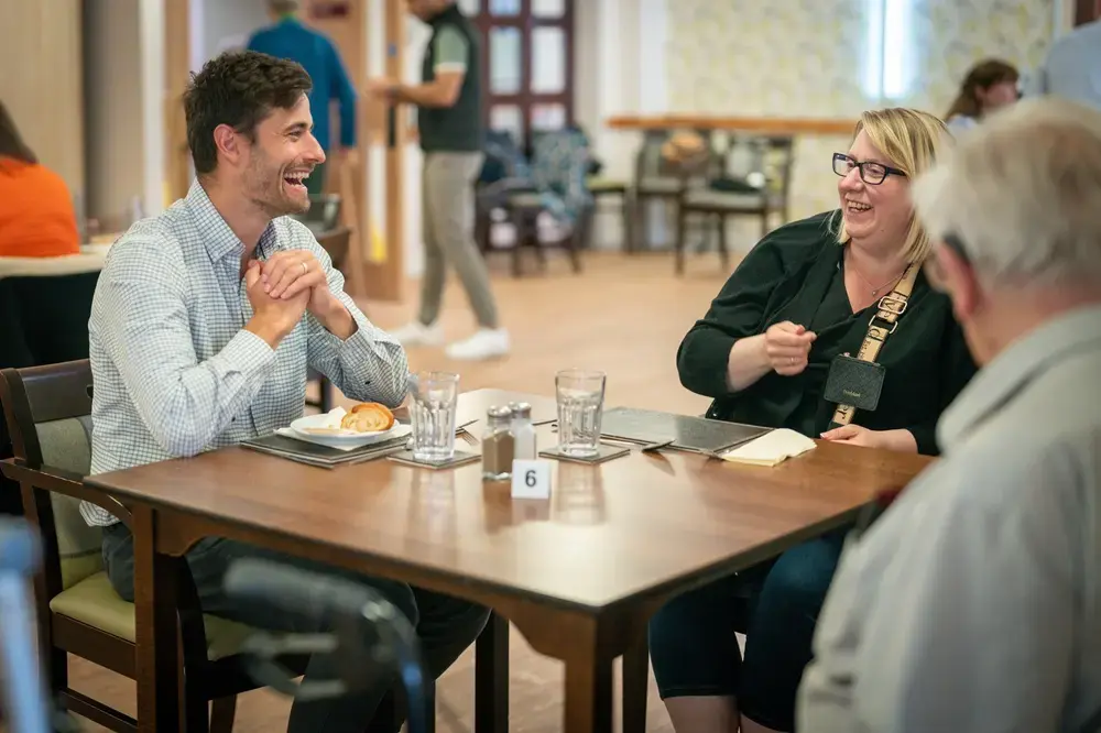 Happy social housing tenants in Wales: Allume Energy CEO Cameron Knox laughing and talking with a couple seated around a dining table in a community room.